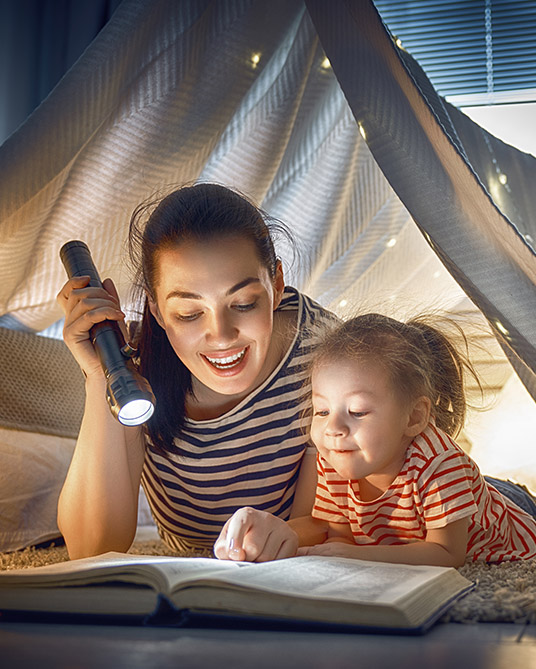 Una madre joven leyendo un libro a la luz de una linterna, junto a su hija, en una carpa 